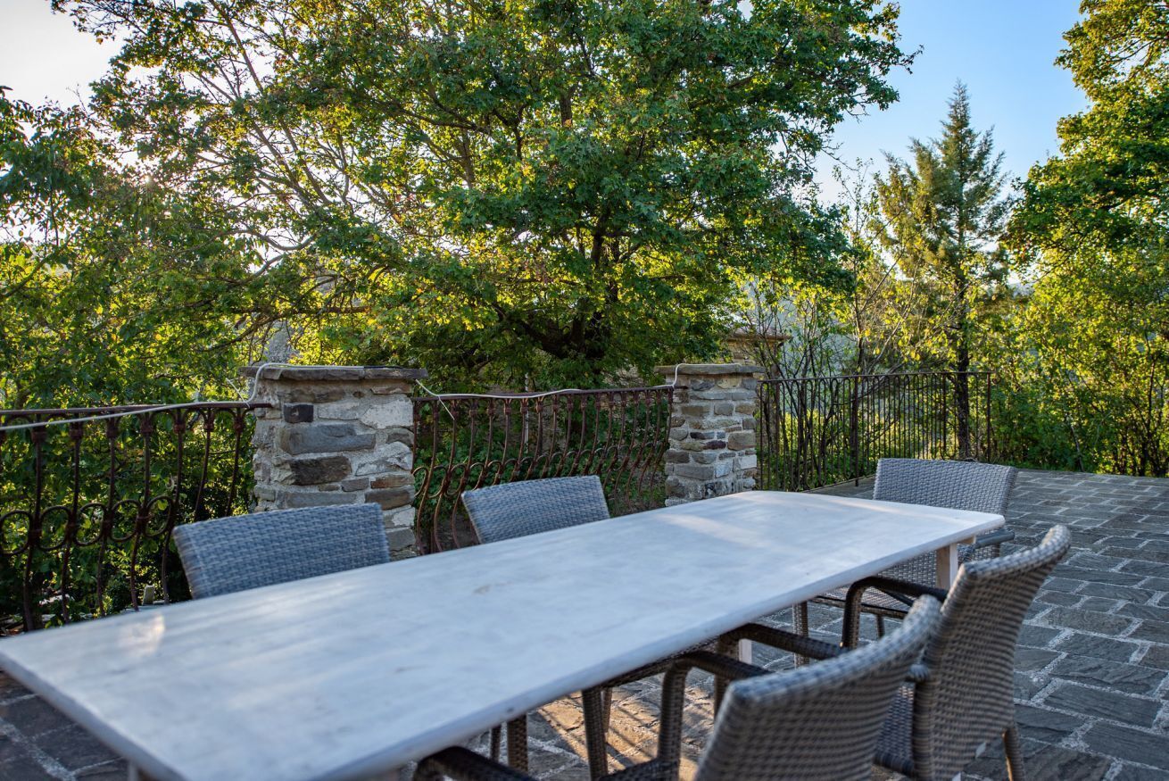 Patio esterno con tavolo in pietra e sedie in vimini, circondato da un muro di pietra e vegetazione.