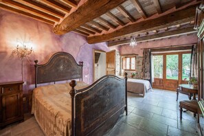 A rustic bedroom with exposed wooden beams, a large bed, a chandelier, and a view of the garden through a door.