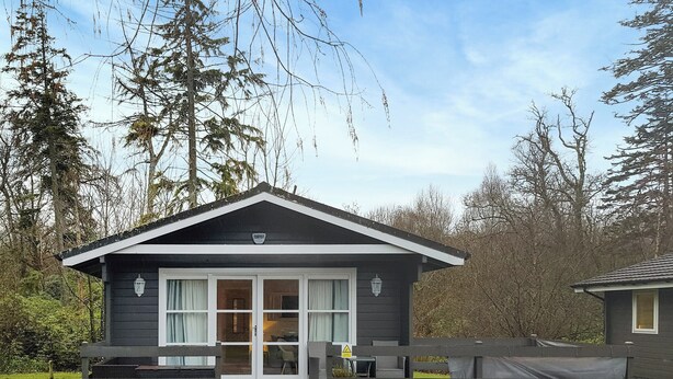 A small, dark-colored cabin with a porch, a sliding glass door, and a small outdoor seating area. - Leven Lodge 1 - UK46042 (Loch Lomond)