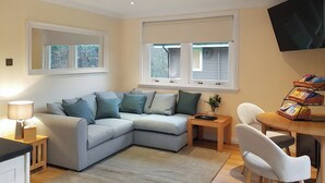 A modern living room with a grey sectional sofa, a small wooden side table, a white armchair, a round wooden table with a magazine rack, and a flat-screen TV mounted on the wall. - Leven Lodge 1 - UK46042 (Loch Lomond)