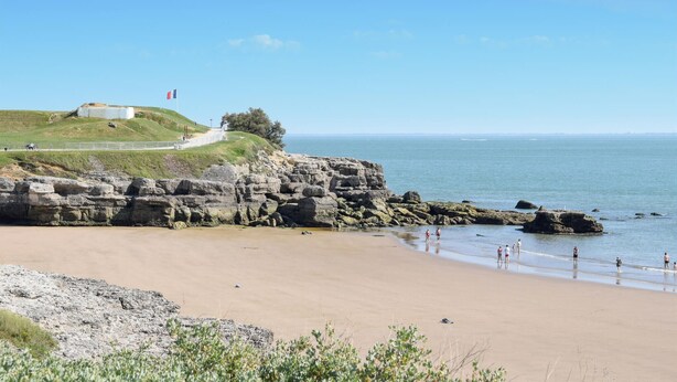 A coastal beach with rocky cliffs, a grassy hill, and a clear blue sky.