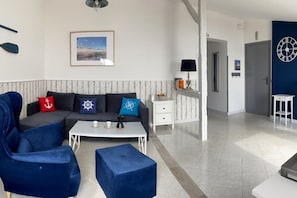 Un salon moderne avec un canapé d’angle, une table basse et des fauteuils bleus. Un tableau encadré est accroché au mur et une horloge est fixée à la porte.