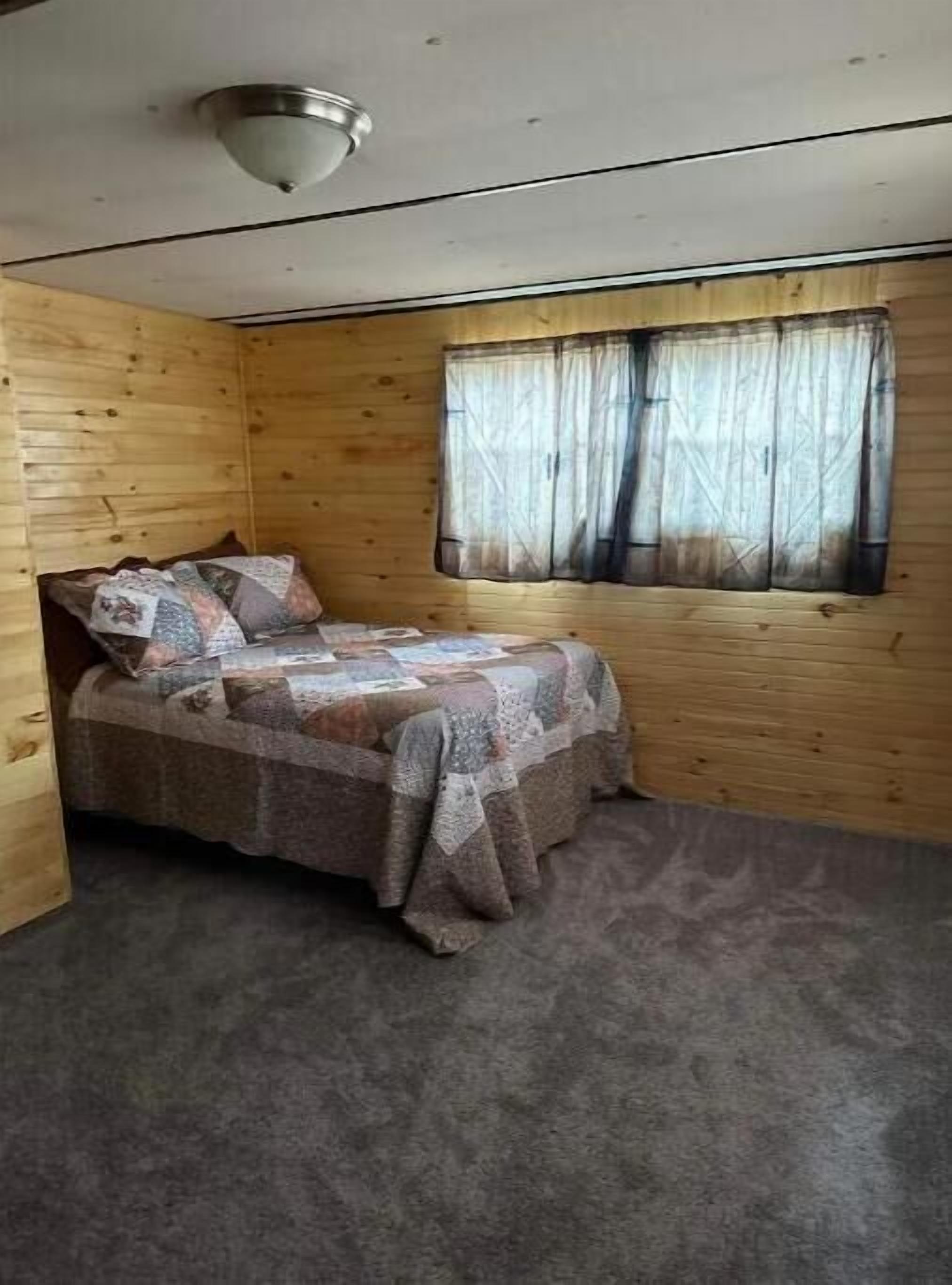 A small, cozy room with a wooden interior, a single bed with a patterned bedspread, a window with sheer curtains, and a ceiling light fixture.