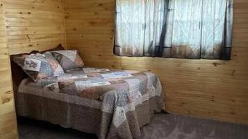 A small, cozy room with a wooden interior, a single bed with a patterned bedspread, a window with sheer curtains, and a ceiling light fixture.