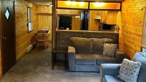 Interior acogedor de una cabaña con fachada de madera, una sala de estar con sofá y una mesa de comedor, y una zona de cocina con armarios y un refrigerador.