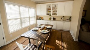 Un comedor con una mesa de madera dispuesta para cuatro personas, sillas blancas y un banco. Hay armarios blancos sobre la mesa con varios objetos en los estantes.