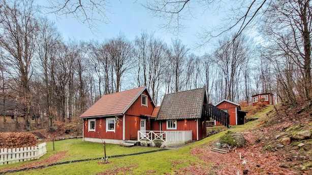 A red wooden house with a sloped roof, a white porch, and a small shed in a forested area. - 1 bedroom awesome home in Ljungbyhed (Ljungbyhed)