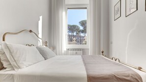 Uma cama bem arrumada com cabeceira, janela com vista para árvores e edifícios, e quadros na parede.