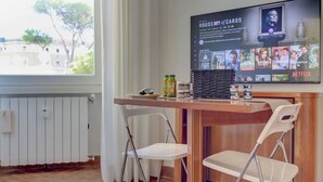Um quarto com uma mesa de madeira, duas cadeiras brancas, uma TV exibindo 'House of Cards' e uma janela com vista para edifícios e árvores.