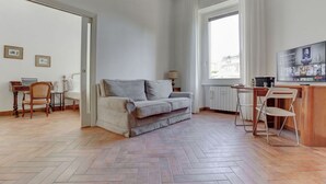 Uma sala de estar moderna com um sofá cinzento, uma mesa de jantar de madeira e uma cozinha com bancada branca e cadeiras.