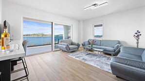 A modern living room with a large sliding glass door leading to a balcony with a view of the sea. There is a sofa, a coffee table, and a TV on a stand.