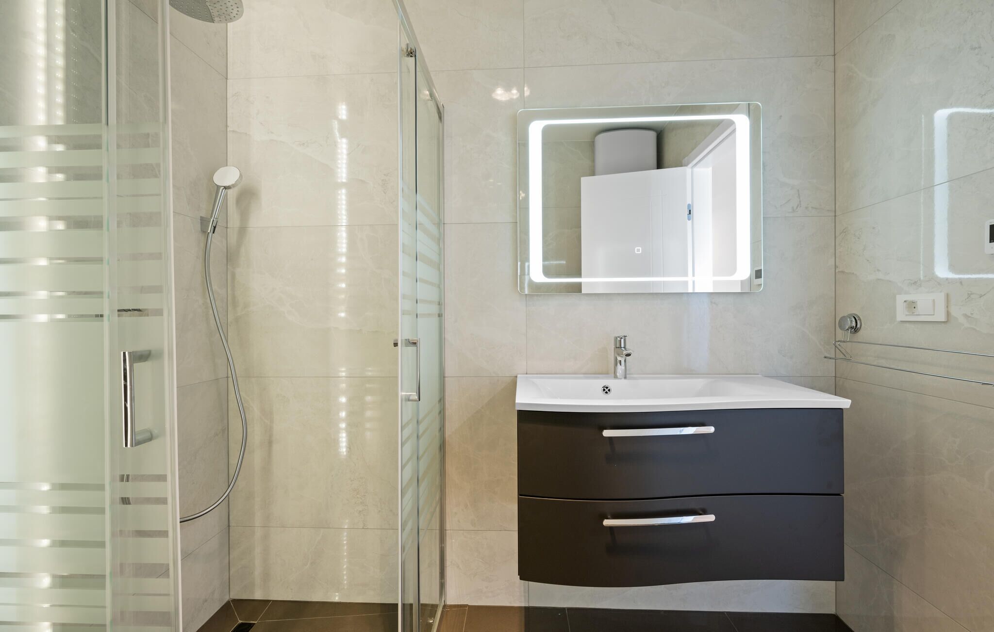 Une salle de bain moderne dotée d’une douche à parois de verre, d’un lavabo blanc avec un meuble noir et d’un miroir avec éclairage intégré.