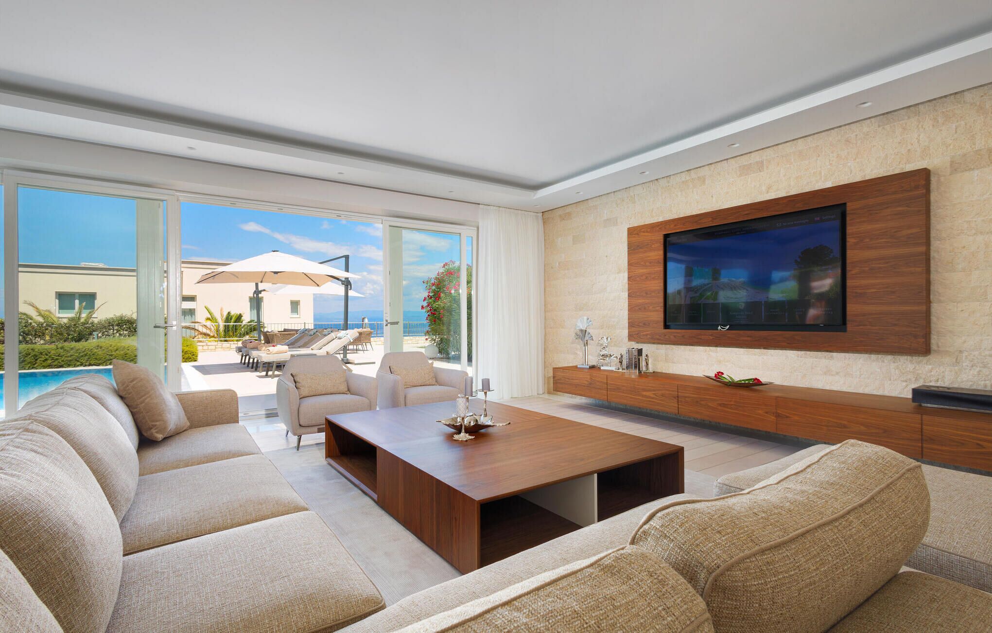 A modern living room with a large TV, comfortable seating, and a view of the pool and outdoors.
