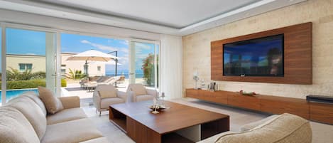 A modern living room with a large TV, comfortable seating, and a view of the pool and outdoors.