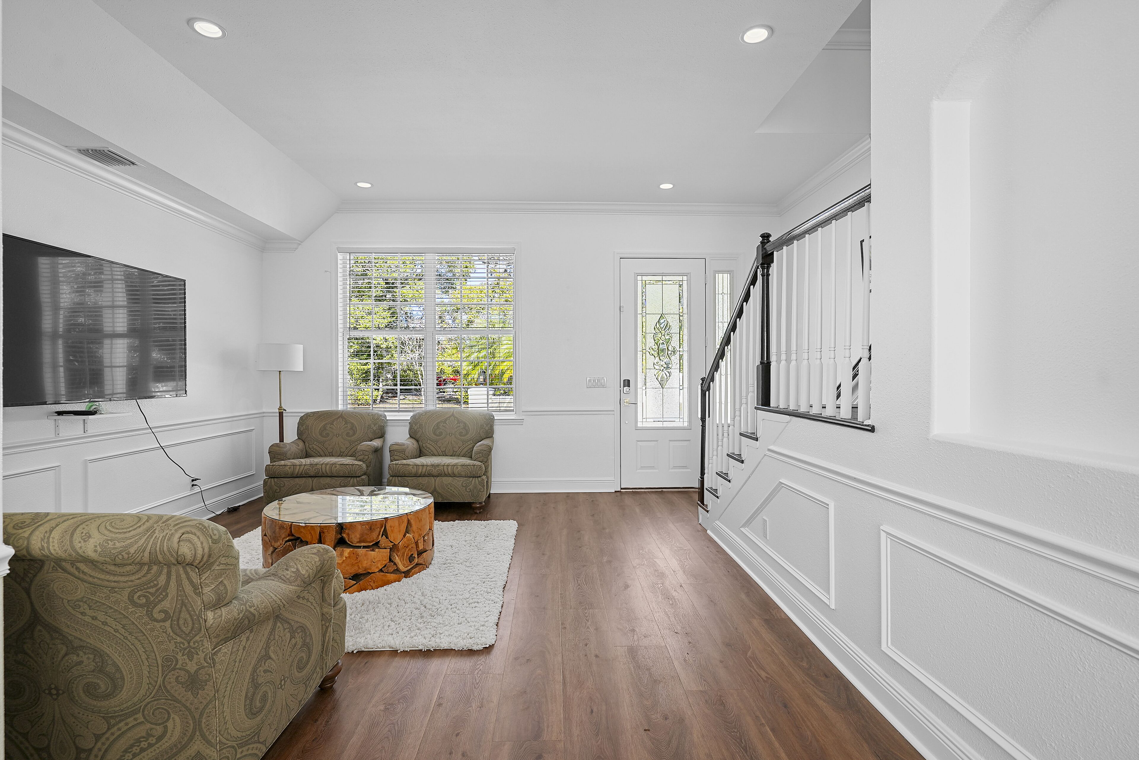 A spacious living room with hardwood flooring, a large flat-screen TV, two patterned armchairs, a wooden coffee table, and a white rug.