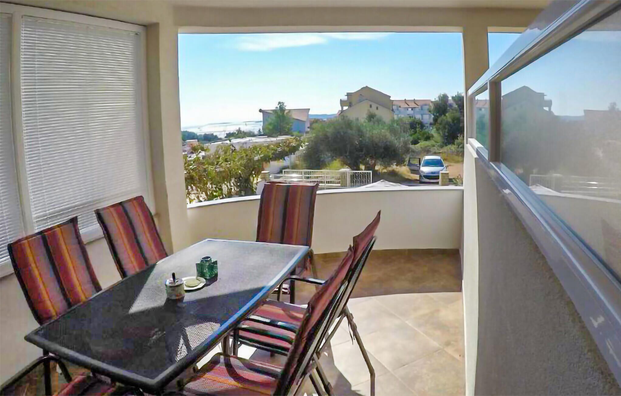 A balcony with a table and chairs, a view of the street and buildings, and a car parked outside.
