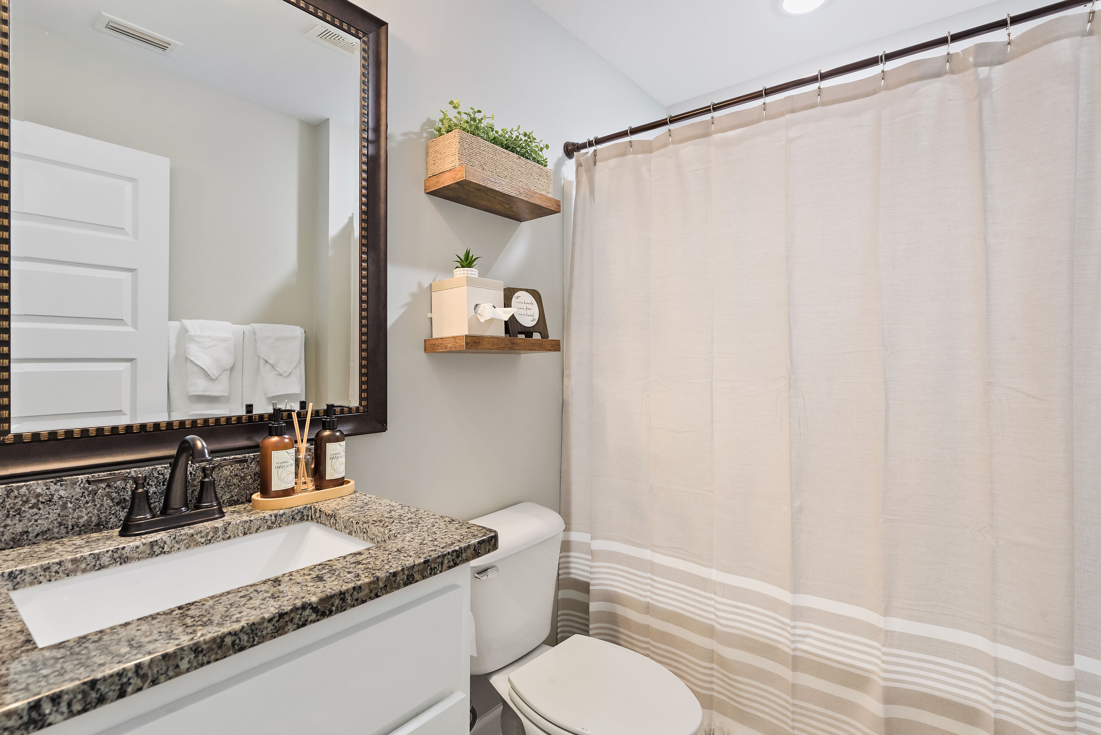 Un bagno con lavabo su piano in granito, un water bianco, tenda della doccia e mensole in legno con articoli da toilette.