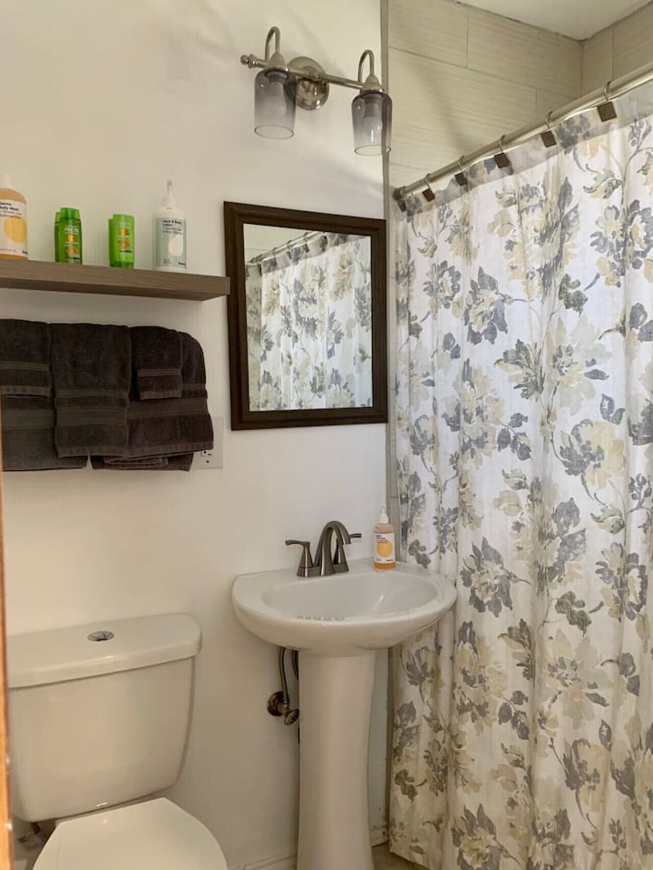 A bathroom with a toilet, sink, and shower curtain. There are toiletries on a shelf above the sink.