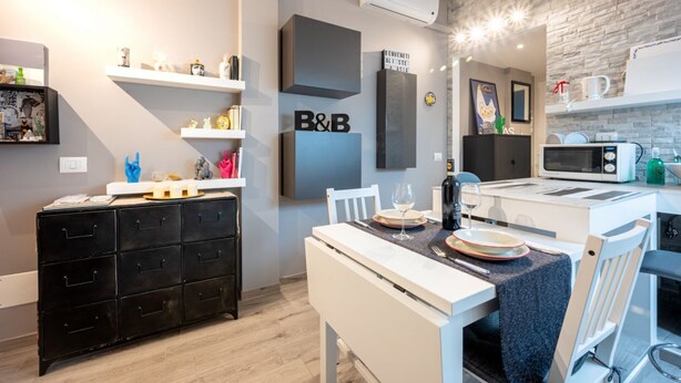 A modern kitchen with a dining area, featuring a black chest of drawers, white chairs, a black and white B&B sign, and a kitchen counter with a microwave. - Comfortable attic located on the third and last floor of a building without a lift.It comfortably accommodates two people in 40 m2 of space with a double bedroom.Small building with a green space and ample free parking in front of the house, near the cen (Parma)