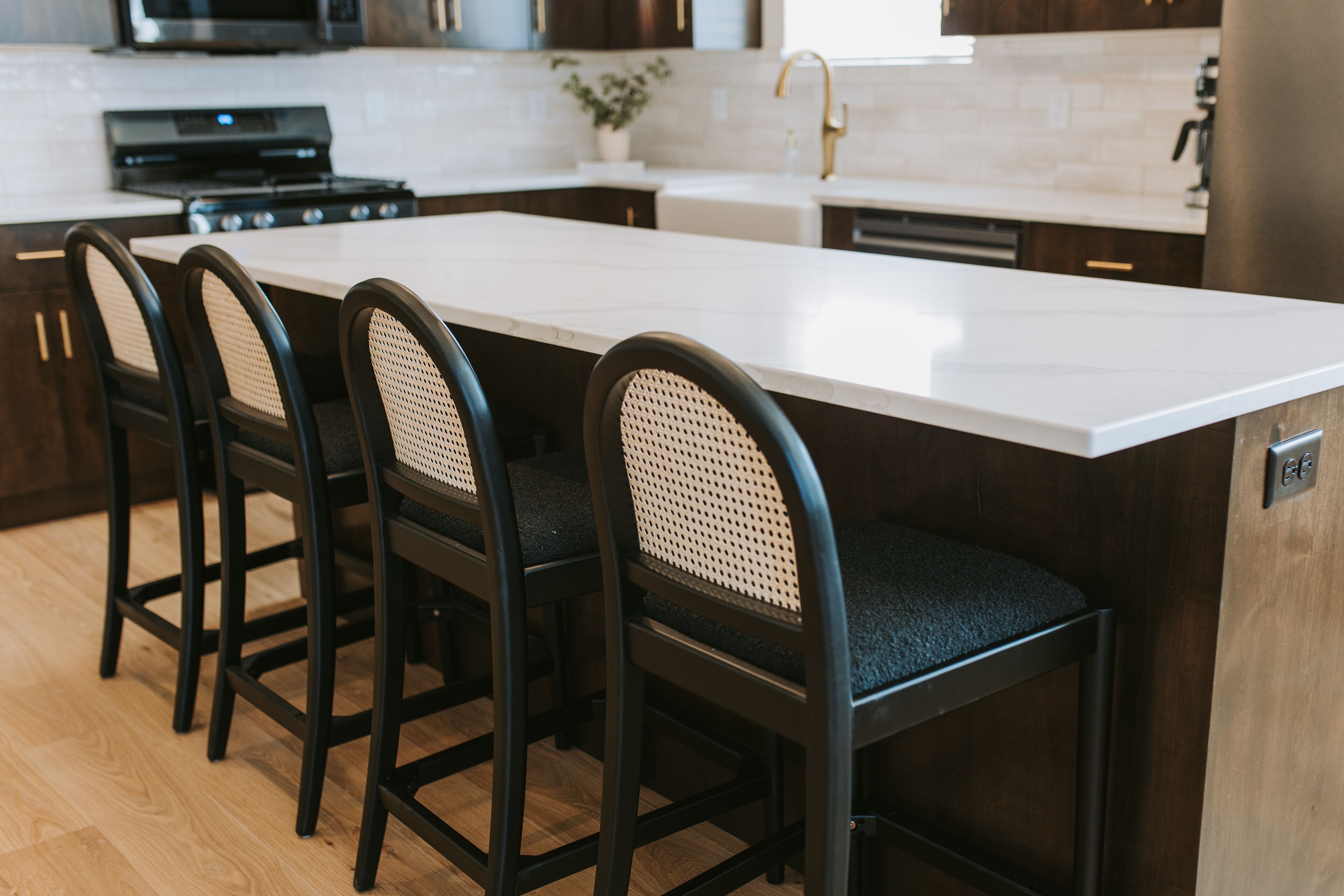 A kitchen with a white countertop, dark wooden cabinets, and three black bar stools with woven seats.