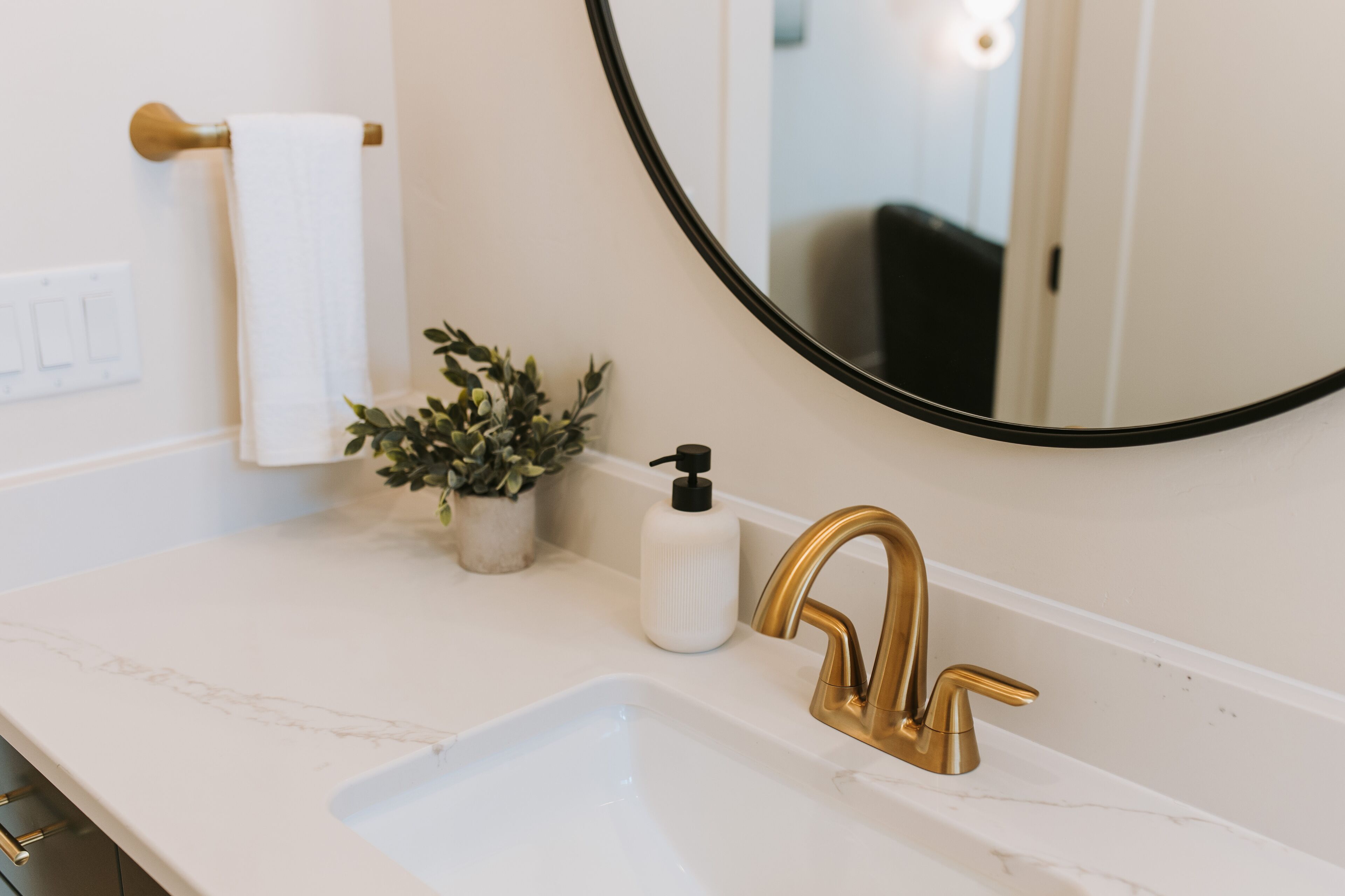 A bathroom with a white marble sink, gold faucet, a potted plant, a white bottle, and a towel hanging on a towel rack.
