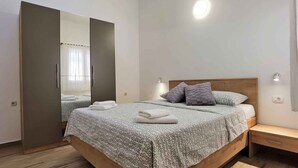 Ein ordentlich bezogenes Bett mit einem gemusterten Überwurf, einem hölzernen Kopfteil und zwei Kissen. Ein Nachttisch mit Lampe und Schublade steht daneben. Auf der linken Seite des Bettes befindet sich ein großer Spiegel, der einen Teil des Raumes reflektiert.