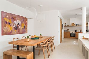 A modern kitchen with wooden cabinets, a white countertop, and a dining area with a wooden table and chairs. - Villa Estrella - Canutells (Es Canutells)