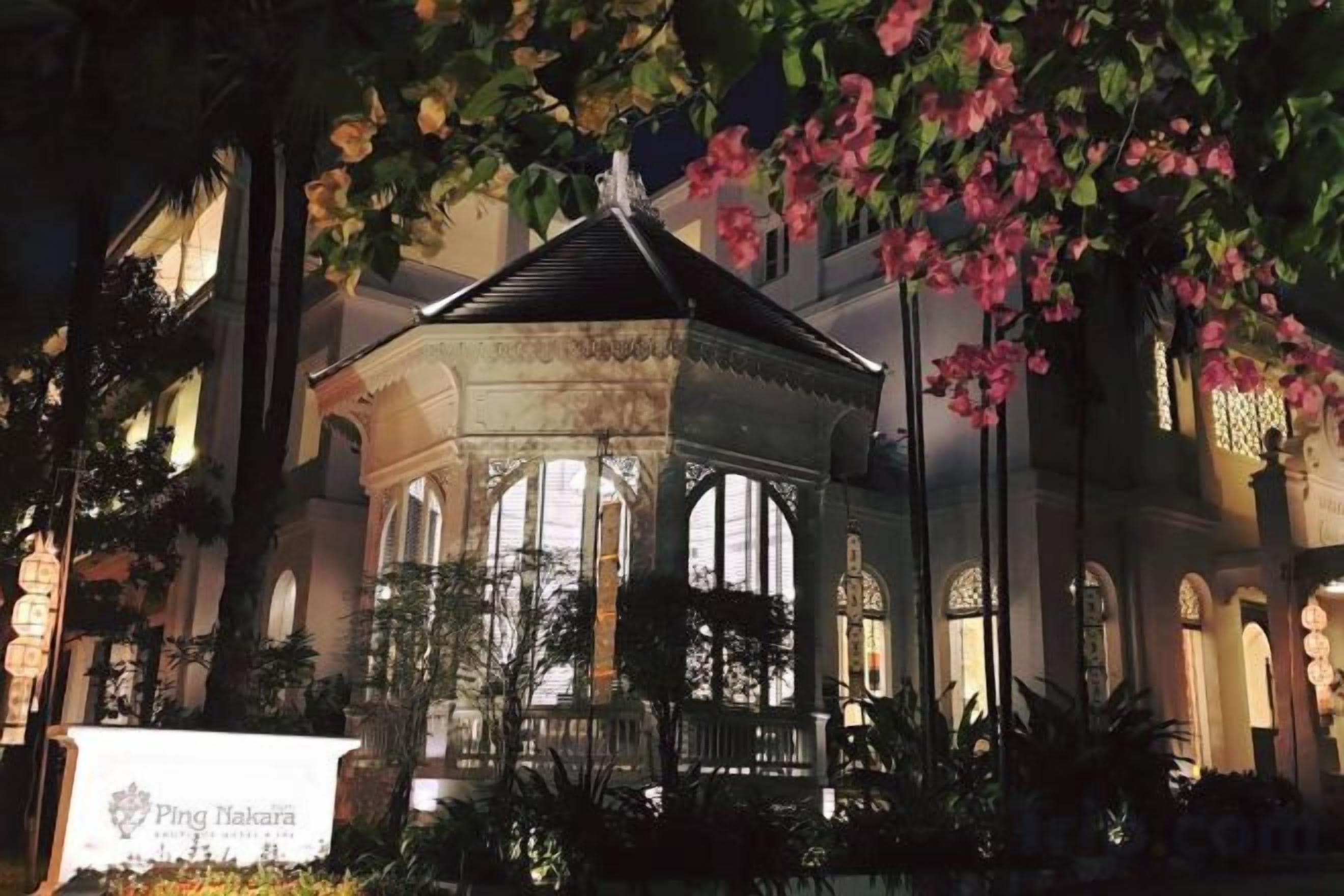 A gazebo with a dome roof, surrounded by lush greenery and a building with arched windows in the background.