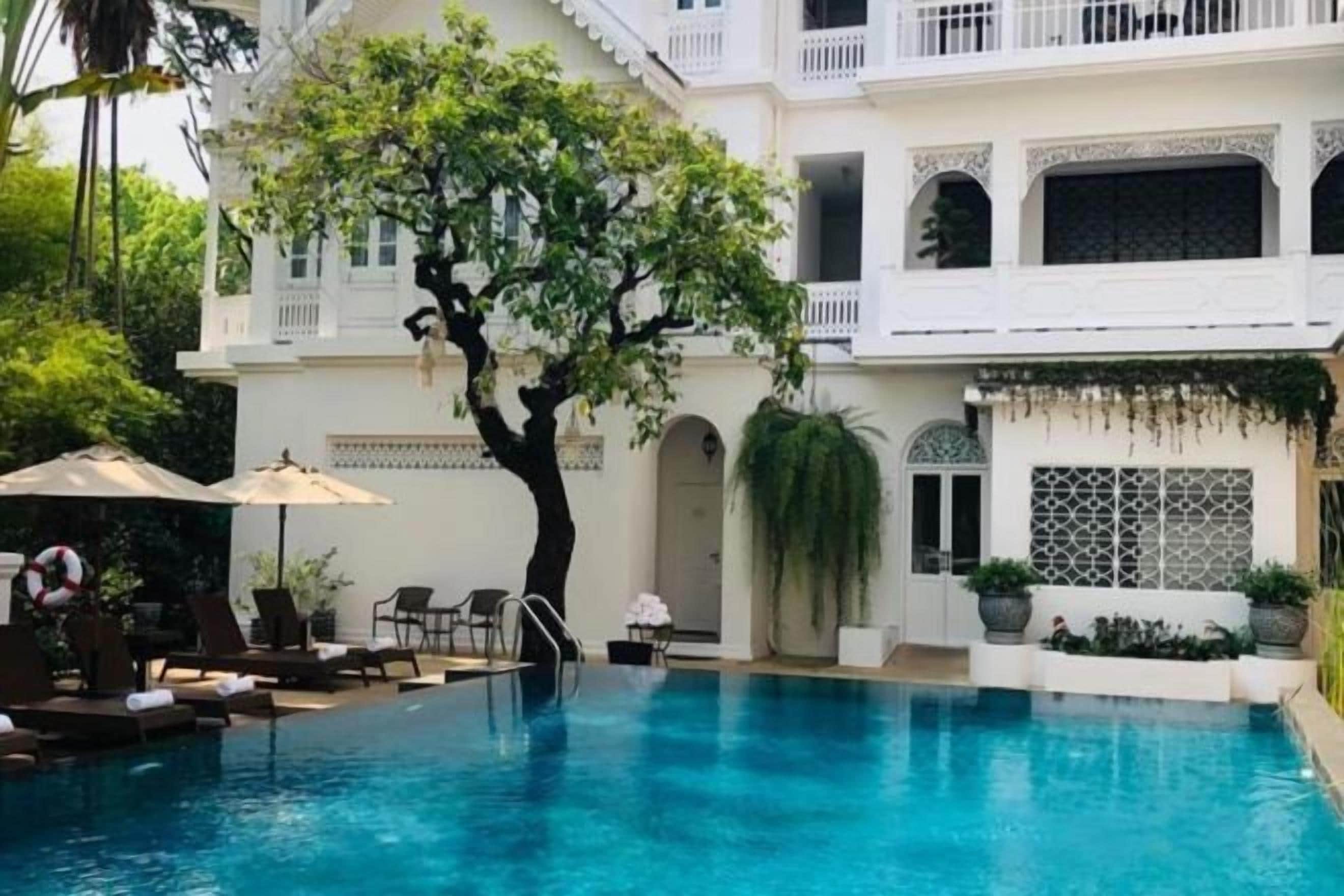A pool area with a tree, lounge chairs, and a white building with balconies.