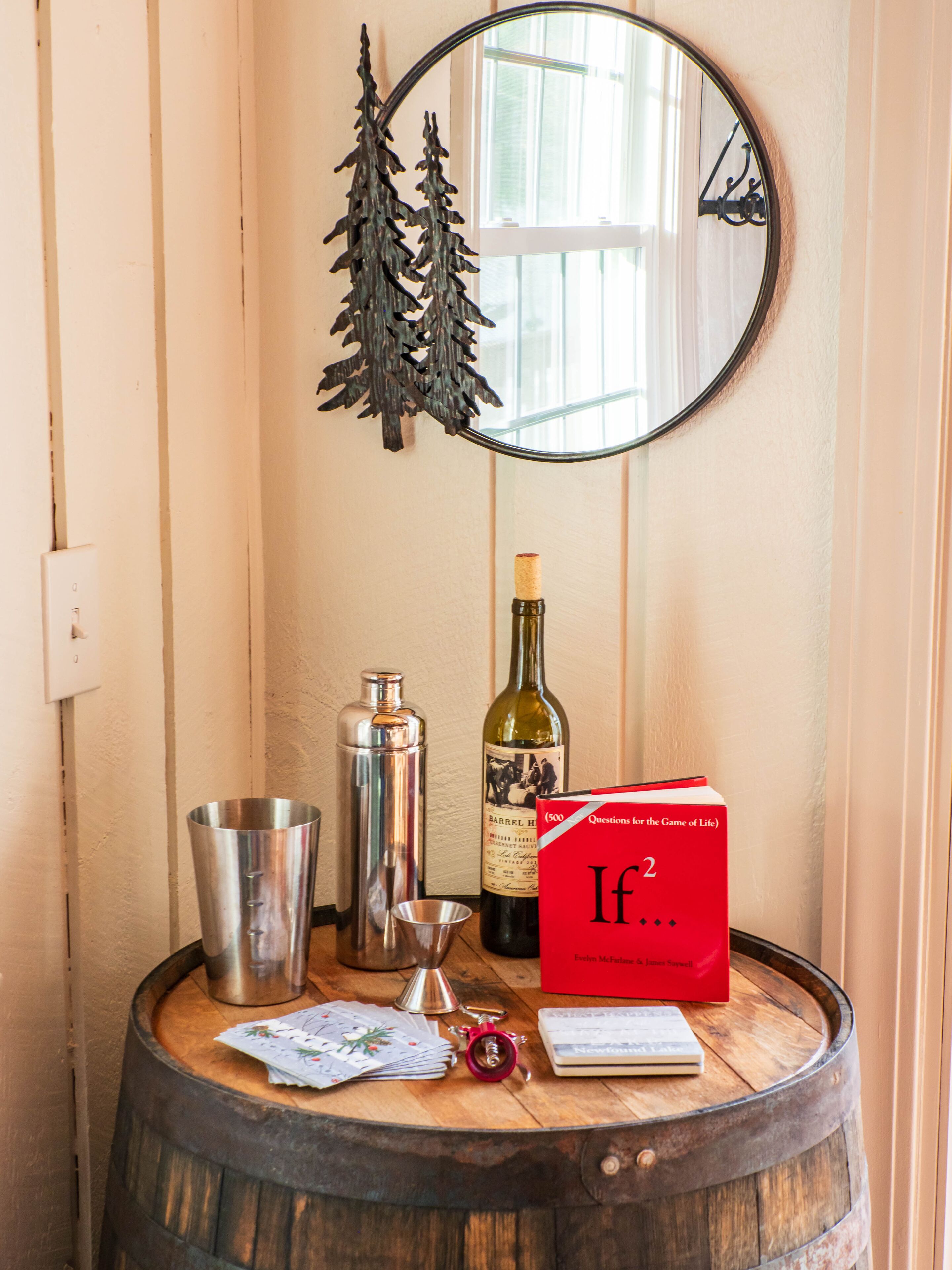 Una mesa de madera con barril, una botella de vino, un coctelero, un libro rojo titulado 'If...', una baraja de cartas y un pequeño objeto rojo.