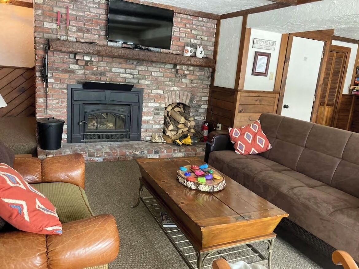 A cozy living room with a brick fireplace, a wooden coffee table, leather sofas, and a television mounted above the fireplace.