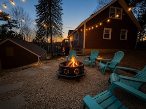 A wooden cabin with a fire pit and outdoor seating, surrounded by trees and string lights.