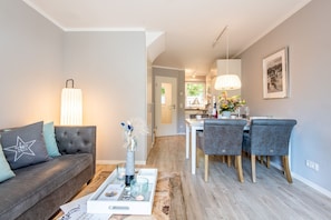 A modern living room with a sofa, coffee table, and dining area with chairs. - Beach House one - Zentral. Familie. Neubau. - Objekt ID. Dbs/12d (Sylt)
