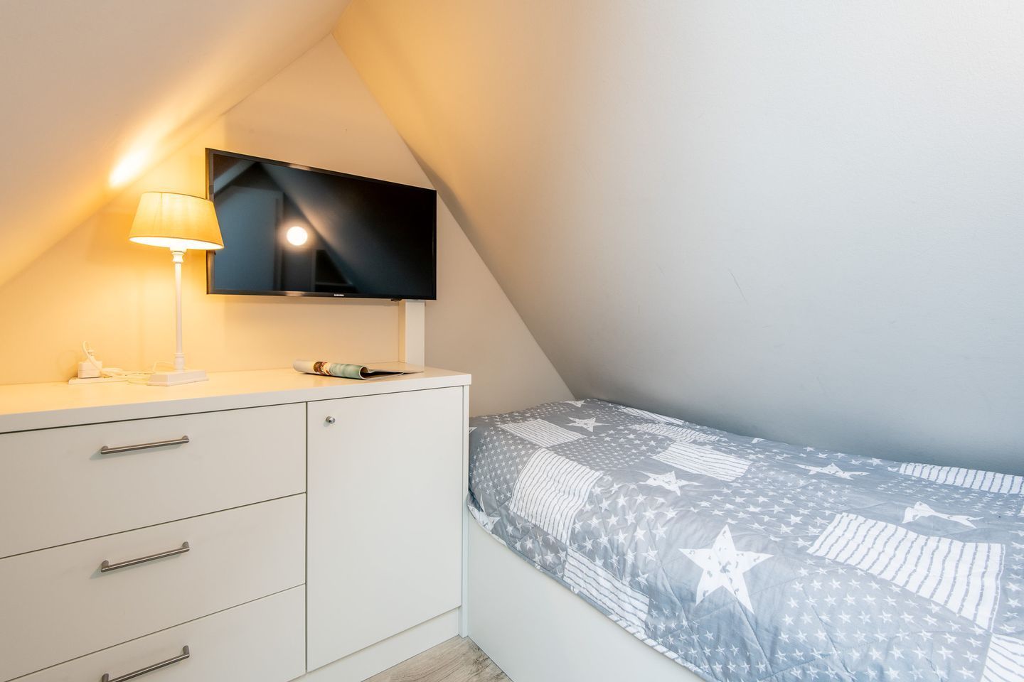 A bedroom with a bed, a nightstand, a lamp, and a flat-screen TV mounted on the wall.