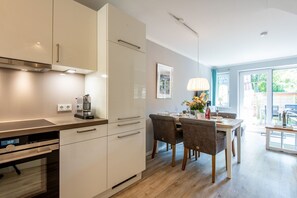 A modern kitchen with built-in appliances, a dining area with a table and chairs, and a large window providing natural light. - Beach House one - Zentral. Familie. Neubau. - Objekt ID. Dbs/12d (Sylt)