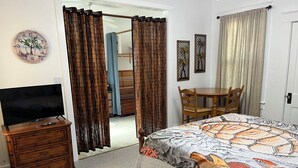 A bedroom with a bed, a TV on a dresser, a chair, a table, and a door with curtains.