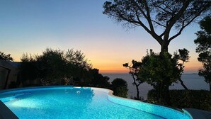 A swimming pool with a curved edge, surrounded by trees and a view of the sea at sunset.