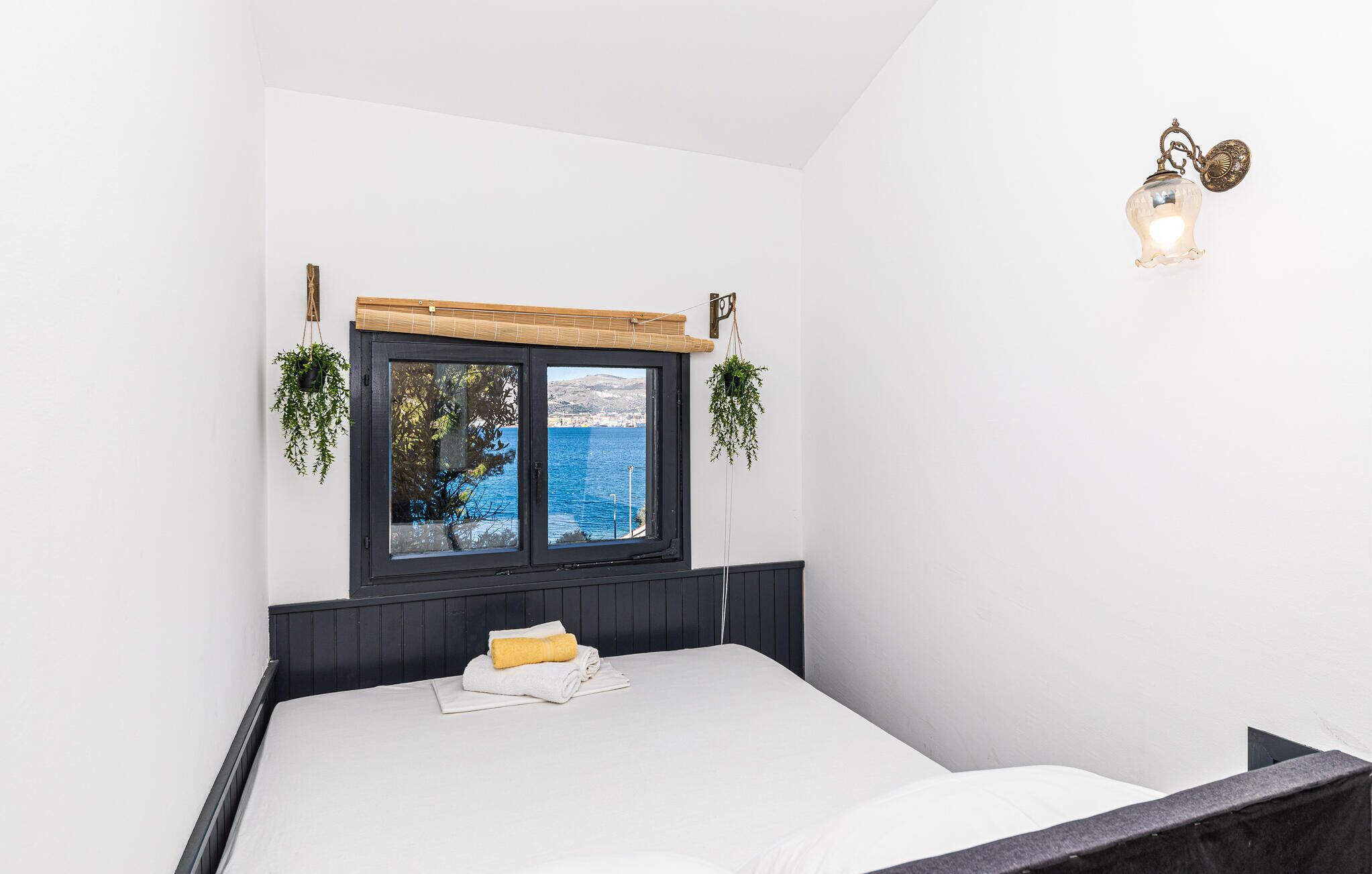 Een slaapkamer met een groot raam met uitzicht op de zee, een bed met een wit matras en een opgerolde handdoek, en een wandlamp.