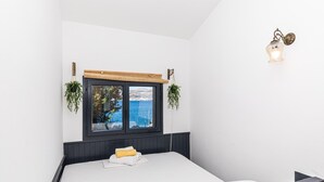 Quarto com uma janela ampla que oferece vista para o mar, uma cama com colchão branco e uma toalha enrolada, e luminária fixada na parede.
