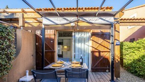 A wooden deck with a table set for four, a covered patio area, and a view of a building with a tiled roof. - Beau Soleil by Interhome (Narbonne-Plage)