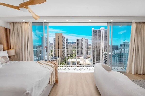 A modern hotel room with a balcony, large windows, and a city view.