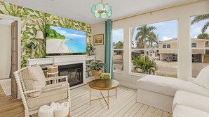 Una sala de estar con televisor, chimenea, sofá y vistas a palmeras fuera.