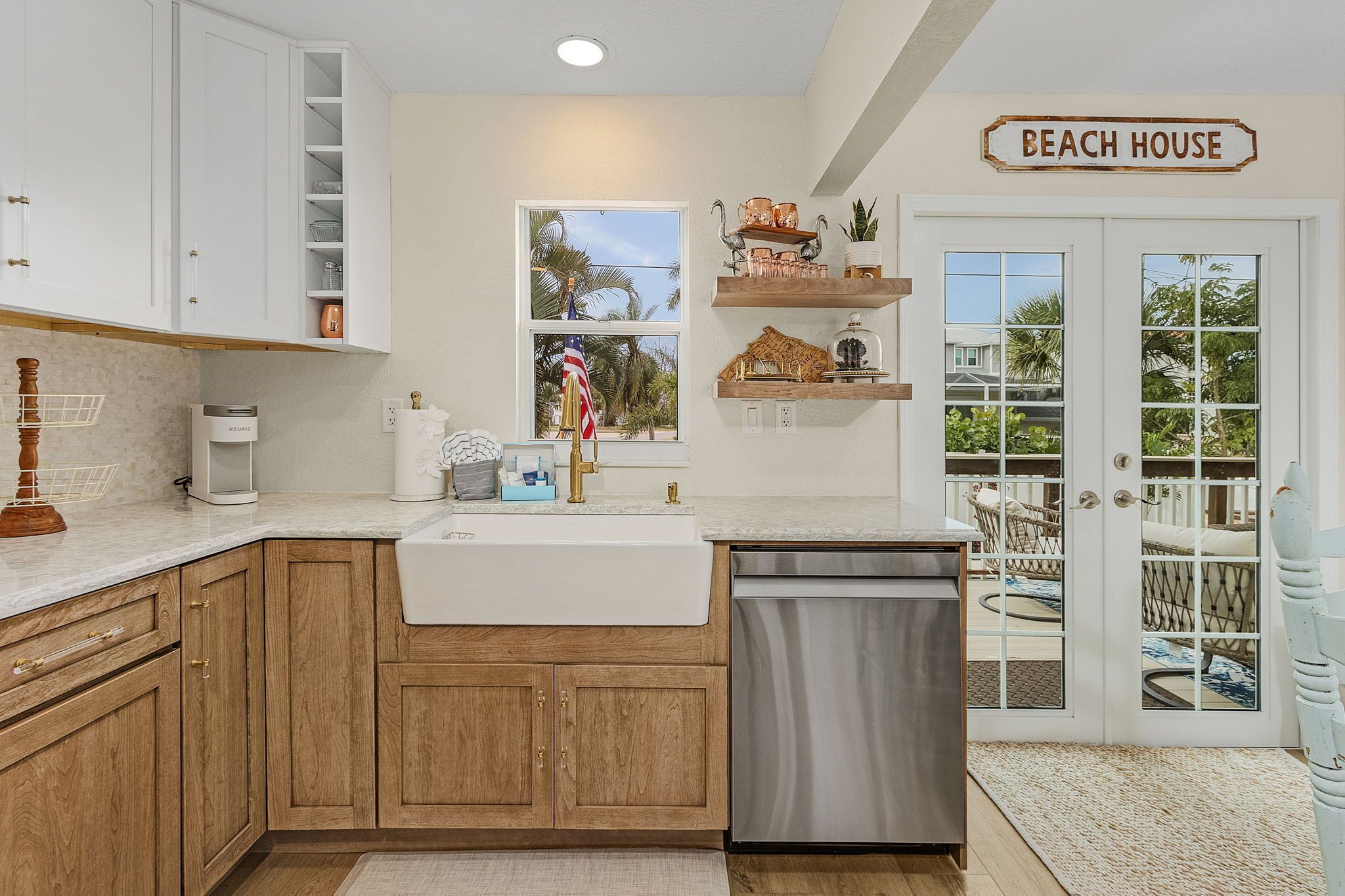 A kitchen with wooden cabinets, a white sink, and a stainless steel dishwasher. There is a window with a view of palm trees and a sign that reads "BEACH HOUSE".