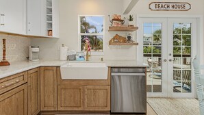 Cocina con armarios de madera, un lavamanos blanco y un lavavajillas de acero inoxidable. Se ve una ventana con vistas a palmeras y un letrero que dice 'BEACH HOUSE'.