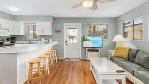 Eine moderne Küche mit weißen Schränken, einer Bar mit Hockern, einem Deckenventilator und einem Fernseher in einem Wohnbereich mit einer Couch und einer Lampe.