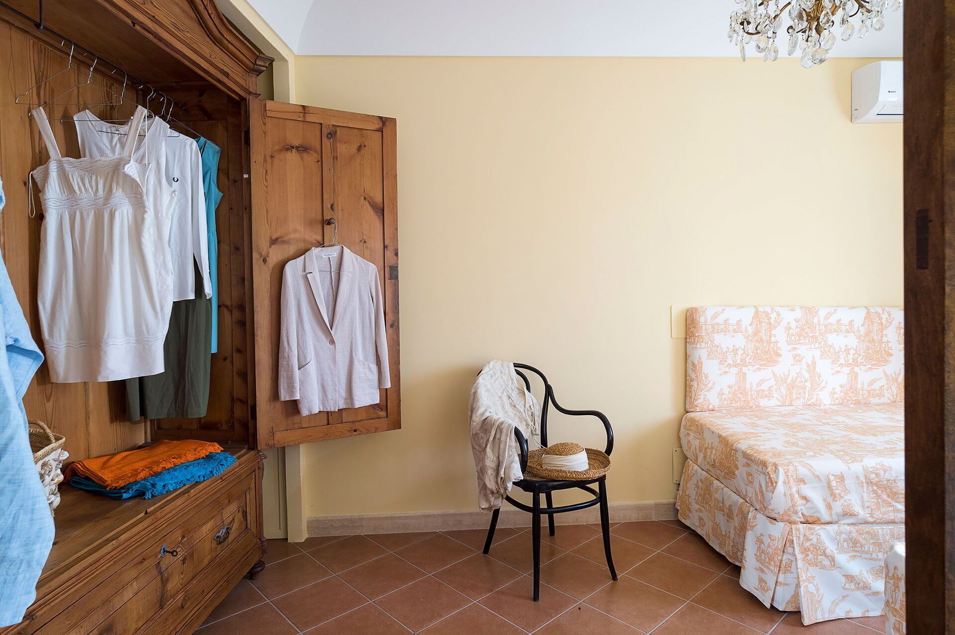 Un dormitorio con un armario de madera, una silla con una cesta tejida, una cama con colcha estampada y una lámpara colgante.