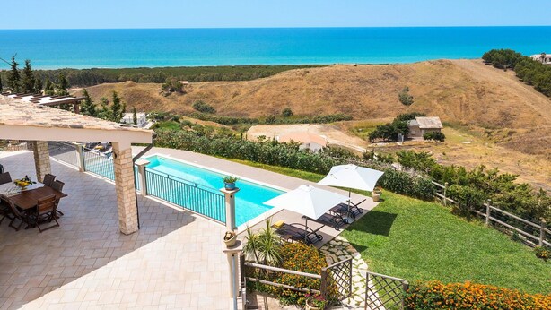 Area piscina con vista sul mare, patio con tavolo e sedie, e paesaggio con prato.