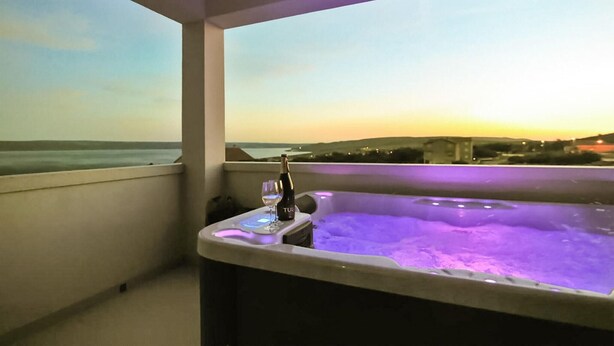 A balcony with a hot tub and a bottle of wine, overlooking a body of water and a distant landscape during sunset.