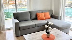 Un salon moderne avec un canapé gris, des coussins orange, une table basse en bois et une vue sur un ensemble d’immeubles à travers de grandes fenêtres.
