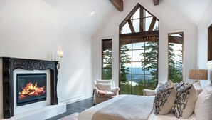 Un dormitorio con chimenea, ventanas amplias, cama con almohadas estampadas y vistas a árboles.